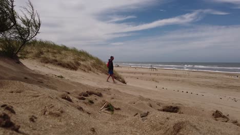 Zoom-in-shot-of-man-walking-between-sand-dunes-to-sandy-beach-during-windy-day---Mar-de-las-Pampas,Argentina