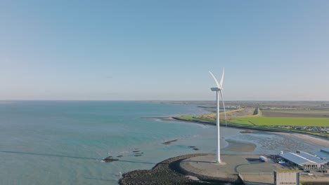 Aerial-slow-motion-shot-of-a-wind-turbine-in-a-rural,-coastal-area-in-the-Netherlands-against-a-blue-sky-on-a-beautiful-sunny-day