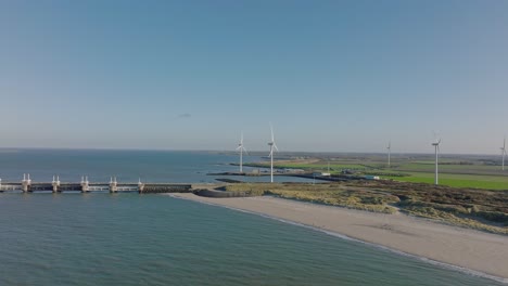 Aerial-slow-motion-shot-of-wind-turbines-and-the-Eastern-Scheldt-storm-surge-barrier-in-a-rural,-coastal-area-in-the-Netherlands-against-a-blue-sky-on-a-sunny-day