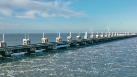 Aerial-slow-motion-shot-of-an-open-Eastern-Scheldt-storm-surge-barrier-and-wind-turbines-in-the-distance-in-Zeeland,-the-Netherlands-on-a-beautiful-sunny-day-with-a-blue-sky