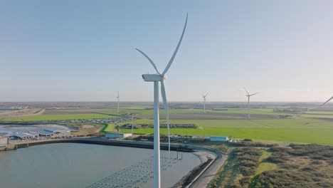 Aerial-slow-motion-shot-of-wind-turbines-and-roads-in-a-rural,-coastal-area-in-the-Netherlands-against-a-blue-sky-on-a-beautiful-sunny-day