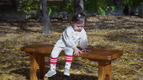 Cute-little-baby-girl-sitting-on-wooden-bench-and-playing-with-autumn-leaves-then-throw-them-on-a-ground-in-Autumn-Park