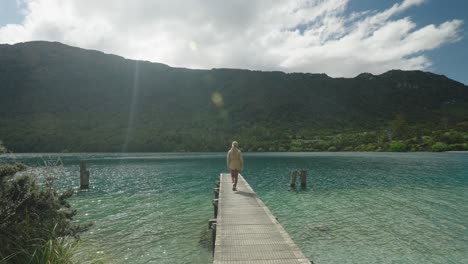 Woman-walking-on-wooden-jetty-at-pristine-blue-cove-with-mountains,-New-Zealand