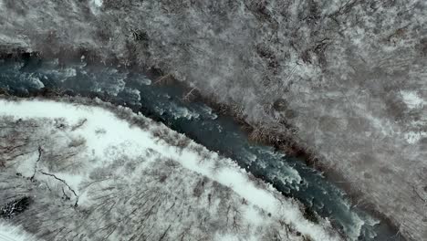 Beautiful-winter-river-flowing-down-snowy-mountainside,-lowering-spinning-aerial