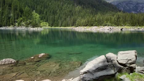 Rocky-beach-of-the-Obernberger-Lake-in-Tyrol-with-turquoise-colored-water-and-a-forest-in-the-background