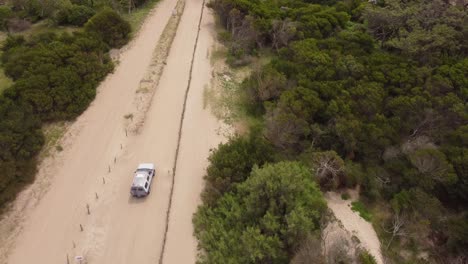 Aerial-tracking-shot-of-white-4x4-wheel-vehicle-driving-on-sandy-beach-path-between-forest-at-sunlight