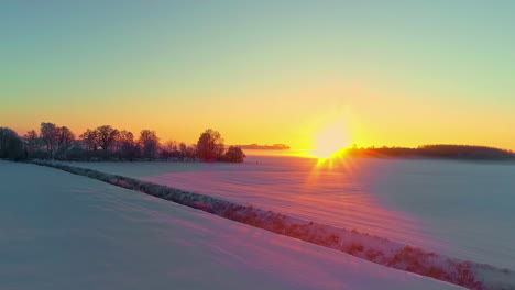 The-brilliant-sunset-reflects-off-fields-of-snow-on-a-cold,-winter-evening---pull-back-aerial-flyover
