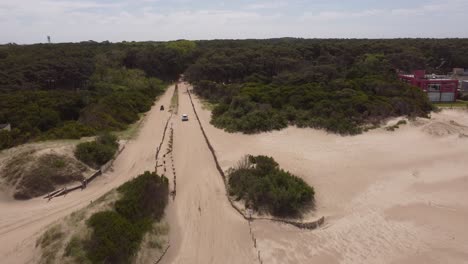 Aerial-view-of-vehicle-driving-on-sandy-path-between-forest-during-sunny-day-at-beach