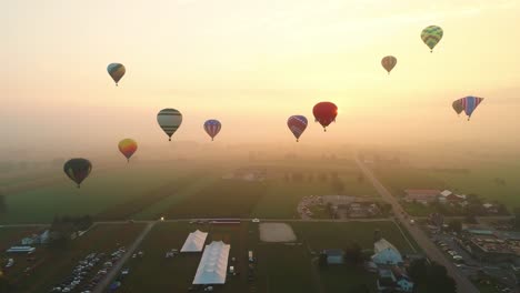 An-Aerial-View-of-Multiple-Hot-Air-Balloons-Rising-into-the-Early-Mist-During-a-Festival-With-Crowds-Watching,-on-a-Summer-Day