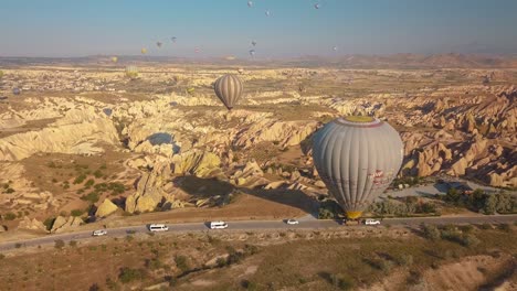 Flying-over-mountains-and-fairy-chimneys-of-Goreme-in-red-valley,-hot-air-balloon-being-transported-on-road-below