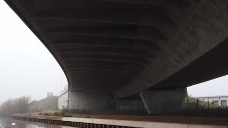 Underneath-large-curved-concrete-motorway-infrastructure-tilt-down-to-misty-sinister-canal-overpass-landscape
