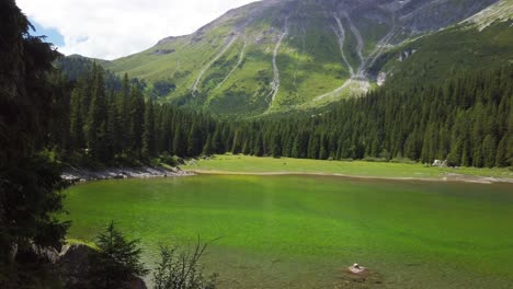 Beach-with-green-colored-water-of-the-Obernberger-Lake-in-Tyrol-,-with-mountains-and-a-forest-in-the-background