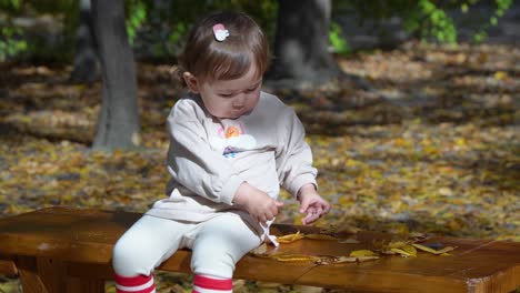 Süßes-Kleines-Mädchen,-Das-Auf-Einer-Holzbank-Sitzt-Und-Mit-Herbstblättern-In-Einem-Park-Spielt