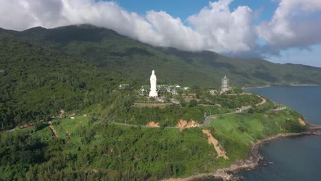 Aerial-drone-flying-towards-tall-Lady-Buddha-statue-and-temples-with-huge-mountains-and-ocean-in-Da-Nang,-Vietnam
