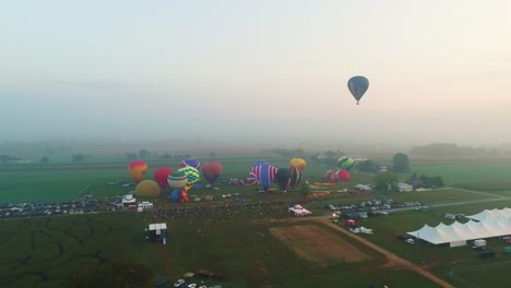 Eine-Luftaufnahme-Mehrerer-Heißluftballons,-Die-Während-Eines-Festivals-Mit-Zuschauern-An-Einem-Sommertag-In-Den-Frühen-Nebel-Aufsteigen