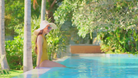Happy-Asian-woman-in-big-sun-hat-and-bikini-at-the-edge-of-swimming-pool