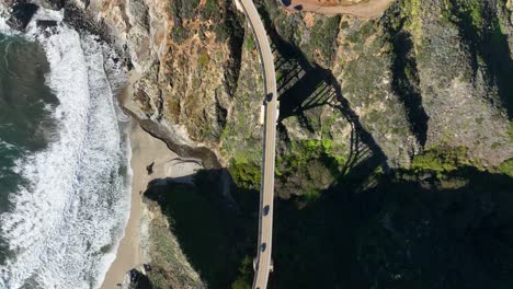 Birds-eye-view-of-Bixby-Bridge-Highway-1-as-waves-crash-on-shore,-Arched-shadow-of-bridge-along-green-hills,-California