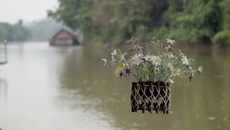 A-close-up-shot-of-a-decorative-flower-pot-suspended-from-the-roof-of-a-bamboo-houseboat,-the-background-filled-with-the-beautiful-scenic-view-of-the-flowing-Khwae-River-in-Kanchanaburi,-Thailand