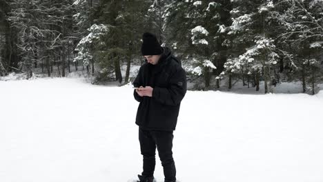 Young-adult-male-texting-on-smartphone-in-a-cold-snowy-winter-pine-forest
