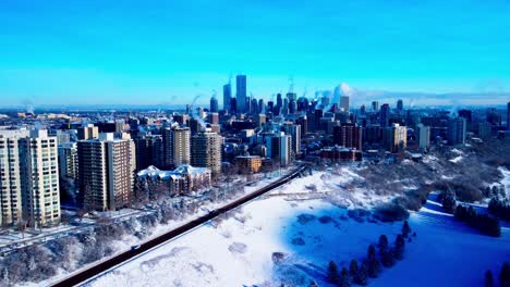 Winter-aerial-drone-hold-over-snow-covered-trees-and-valley-edge-of-the-North-West-view-of-downtown-capital-city-of-Edmonton-Alberta-with-the-tallest-buildings-protruding-over-the-blue-clear-horizon