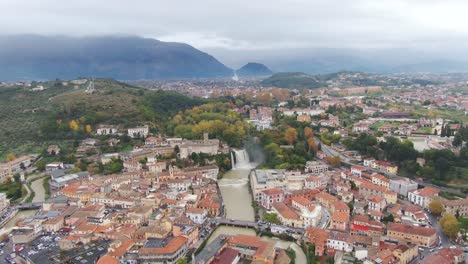 Picturesque-view-of-downtown-Lazio,-Isola-del-Liri-waterfall,-fortified-palace-and-mountains-in-background-on-cloudy-day,-Italy,-aerial
