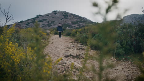 Man-with-camera-walks-on-a-rocky-trail-among-bushes-and-wild-flowers