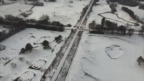 Car-Driving-On-A-Snow-Covered-Road-Surrounded-By-A-Golf-Course-During-Winter