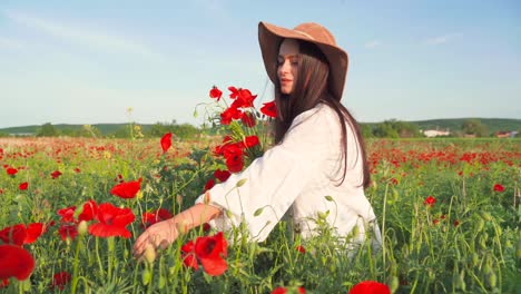 Caucasian-woman-wearing-hat-picks-red-poppies-and-smells-flowers-in-picturesque-field,-handheld-close-up-pan