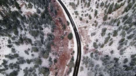 Drone-Aerial-View-Of-Car-Driving-On-Black-Snow-Covered-Winter-Road-Surrounded-By-Pine-Tree-Woodland-Hills-Valley-Near-Kittredge-Evergreen-Colorado