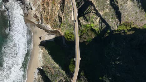 Top-down-view-following-car-along-highway-1-coastline,-Waves-and-seashore-below,-Bixby-Bridge-Big-Sur,-California