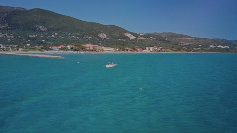 Wide-aerial-as-small-boat-bobs-in-blue-water-near-shoreline-of-Greek-island