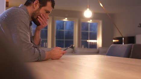 Man-sits-at-table-reading-text-message-and-pushes-his-phone-away-in-disbelief