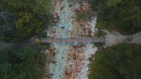 Guy-in-yellow-rain-coat-crossing-small-bridge-above-clear-mountain-water-river,-top-down