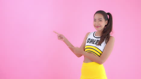 Asian-woman-in-bright-yellow-cheerleader-outfit-against-pink-background-points-with-both-hands-toward-empty-copy-space-on-left