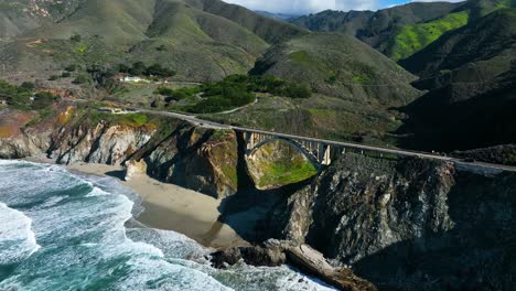 Aerial-cinematic-shot-of-Rocky-Creek-Bridge,-waves-crashing-on-shore-below-against-Green-mountain-range,-Highway-1-California