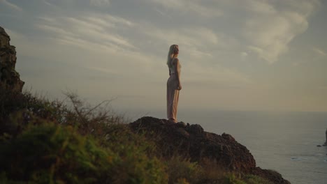 Charming-blond-woman-standing-on-cliff-edge-spinning-head-swaying-hair,-morning-sunlight