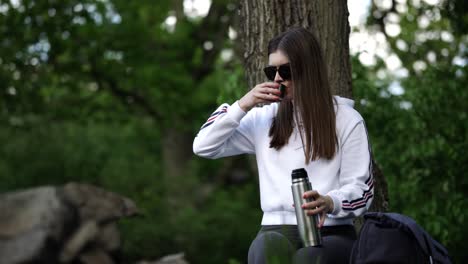 Beautiful-young-caucasian-girl-sitting-in-forest-and-drinking-hot-beverage-from-thermos-bottle