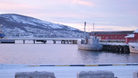 Toma-Panorámica-A-Través-De-Barcos-Y-Barcos-Amarrados-En-El-Muelle-Y-El-Muelle-Durante-La-Noche-Polar-Sobre-El-Círculo-Polar-ártico-En-Narvik,-Norte-De-Noruega-Con-Colinas-Cubiertas-De-Nieve-En-El-Fondo