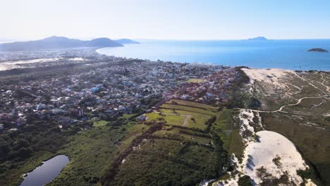Aerial-drone-scene-paradisiacal-beach-florianópolis-seen-from-the-top-seaside-town-near-the-sea-with-mountains-dunes-horizon-buildings-urban-subdivision-on-the-coast-Praia-dos-Ingles
