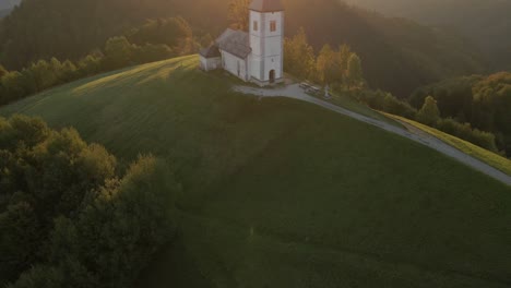 Reveal-shot-of-iconic-St-Primoz-church-on-hill-top-with-bright-morning-sunlight,-aerial