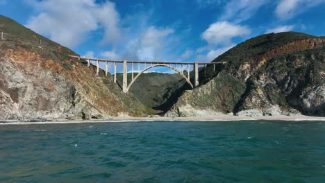 Cinematic-shot-of-Bixby-Bridge-against-green-mountain-range-as-waves-crash-onto-the-shore,-Big-Sur-California