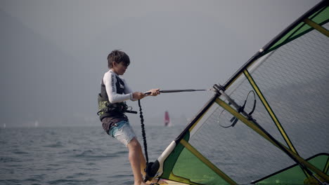 Young-boy-raises-windsurfing-sail-in-ocean,-tracking