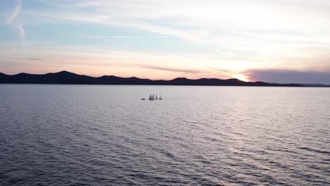 Small-training-sail-boats-in-open-sea-with-silhouette-mountains-during-bright-sunset,-aerial