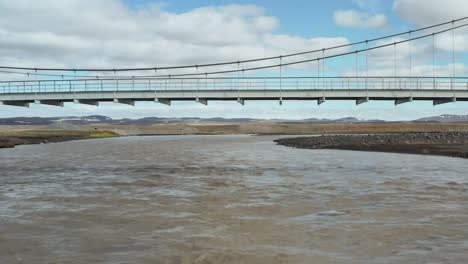 Flying-under-hanging-suspension-bridge-at-glacial-river-in-Iceland,-aerial