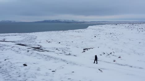 Man-walking-in-rugged-white-snowy-terrain-at-Iceland-coast,-aerial