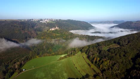 Drone-flight-on-the-countryside-of-Luxembourg-highlighting-a-medieval-castle-on-the-top-of-the-hill,-while-fog-is-slowly-rolling-into-the-valleys