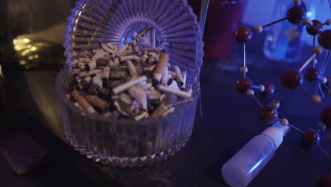 Close-up-of-Smokers-hand-with-lit-cigarette-using-full-ashtray-on-a-table-with-blue-lighting-like-party