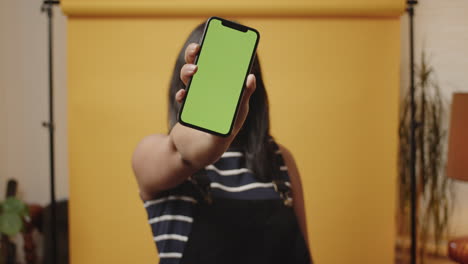 Smiling-young-Asian-woman-holds-a-smartphone-with-a-greenscreen-out-towards-camera-in-a-home-studio