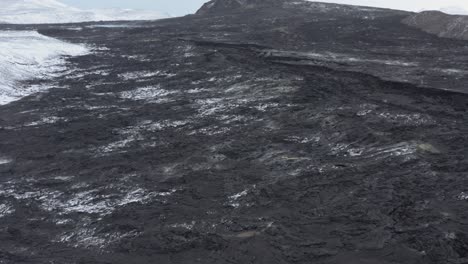 Tourist-sitting-on-snowy-hill-overlooking-black-basalt-lava-field-at-Iceland-volcano,-aerial