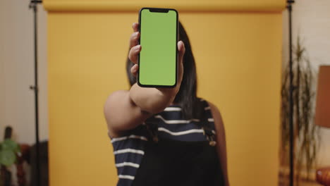 Young-Asian-woman-quickly-shows-her-smarphone-with-a-green-chroma-key-in-a-yellow-studio